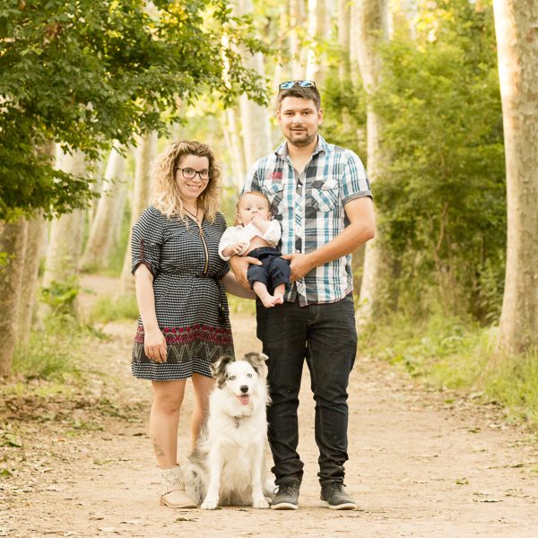 Hombre y mujer de pie en un camino de tierra rodeado de árboles, con un bebé y unPerro border collie blanco y negro